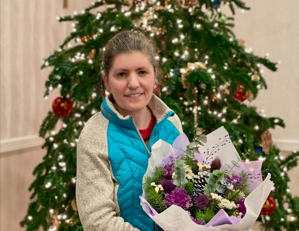 A woman smiles while holding a colorful bouquet of flowers in front of a decorated Christmas tree with lights and ornaments.