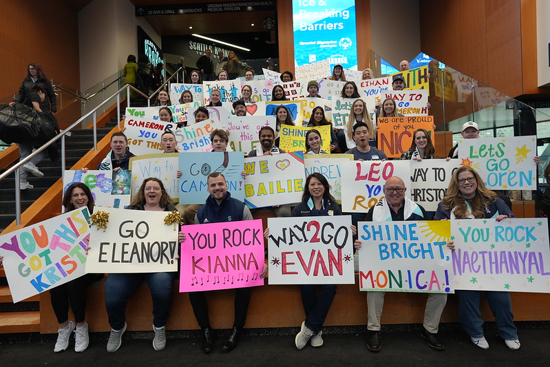 A large, enthusiastic group holds colorful handmade signs cheering on athletes at the Breaking Ice and Breaking Barriers event, gathered on a staircase inside the venue.